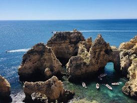 Several boats navigate through turquoise waters around stunning rock formations with arches and cliffs. The horizon shows a vast expanse of calm, deep blue sea, under a clear blue sky.