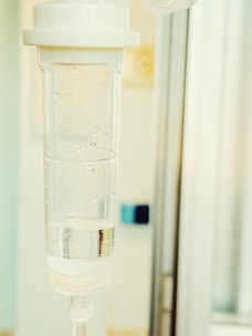 A close-up view of a medical intravenous (IV) drip chamber, partially filled with a clear liquid and several air bubbles. The background appears to be a sterile hospital or clinical setting, characterized by neutral and light-colored surfaces.