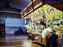 A vibrant carnival stall brightly lit with colorful lights, displaying an array of sweets such as cotton candy and other treats. The scene is set at night in a plaza with a building and white tents in the background. A person stands under an umbrella next to the stall, suggesting light rain.