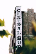 A tall brick building with a white vertical sign reading 'CENTRAL SUP'. The building has a classic industrial look with its red bricks and simple architectural design. Tree leaves partially obscure the lower part of the image, giving a sense of depth and natural elements.