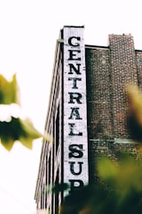 A tall brick building with a white vertical sign reading 'CENTRAL SUP'. The building has a classic industrial look with its red bricks and simple architectural design. Tree leaves partially obscure the lower part of the image, giving a sense of depth and natural elements.