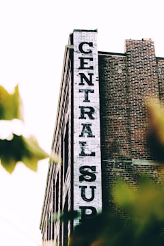 A tall brick building with a white vertical sign reading 'CENTRAL SUP'. The building has a classic industrial look with its red bricks and simple architectural design. Tree leaves partially obscure the lower part of the image, giving a sense of depth and natural elements.