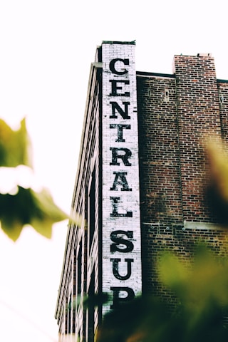 A tall brick building with a white vertical sign reading 'CENTRAL SUP'. The building has a classic industrial look with its red bricks and simple architectural design. Tree leaves partially obscure the lower part of the image, giving a sense of depth and natural elements.