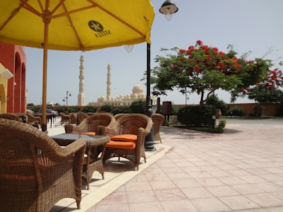 A patio area with wicker chairs and tables shaded by a large yellow umbrella. In the background, there are elegant mosque minarets and domes. A vibrant red-flowering tree and manicured bushes add to the tranquil setting.