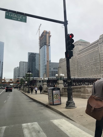 An urban street scene with a tall building under construction, featuring a crane on top. The road is lined with classic-style lamp posts and a few pedestrians are visible on the sidewalk. A traffic light shows red and a street sign reads 'N LASALLE ST 140 W'. A bridge crossing the road carries an elevated train.