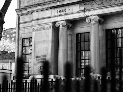 A historical building with tall, classical columns and large windows, displaying architectural details and the engraved year 1848. The sign reads 'Montclare Children's School.' A partially visible bus is in front, and a wrought iron fence is in the foreground.