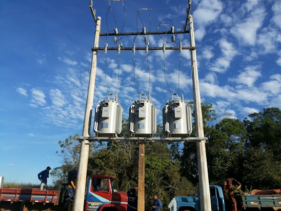 Three electrical transformers are mounted on a pole against a clear blue sky with scattered clouds. There are two trucks parked below, and two people are standing nearby, possibly performing maintenance work. The surrounding area includes green trees and vegetation.