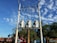 Three electrical transformers are mounted on a pole against a clear blue sky with scattered clouds. There are two trucks parked below, and two people are standing nearby, possibly performing maintenance work. The surrounding area includes green trees and vegetation.