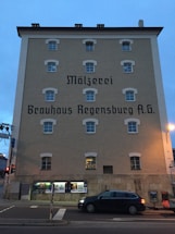 A multi-story building with a beige facade and several windows is prominently displayed. The building features large, stylized text that reads 'Mälzerei Brauhaus Regensburg A.G.' A parked car is visible in front on the street, and various posters are displayed on the ground level of the building. The sky is a dusky blue, indicating the time is likely early evening or late afternoon.