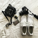 A neatly arranged collection of personal items on a white surface, including a black camera with a strap, white athletic shoes with visible brand text, a small black bag with a noticeable brand logo, a car key fob, and a decorated credit card.
