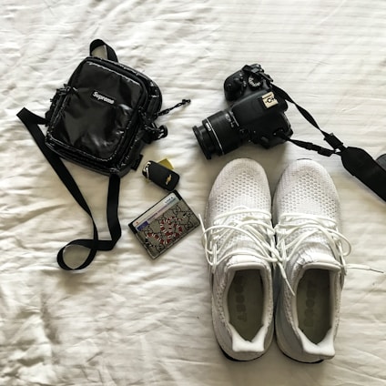 A neatly arranged collection of personal items on a white surface, including a black camera with a strap, white athletic shoes with visible brand text, a small black bag with a noticeable brand logo, a car key fob, and a decorated credit card.