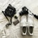 A neatly arranged collection of personal items on a white surface, including a black camera with a strap, white athletic shoes with visible brand text, a small black bag with a noticeable brand logo, a car key fob, and a decorated credit card.