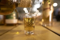 A shot glass filled with a golden liquid is being poured from a woven-covered bottle. The glass is placed on a wooden surface and has a logo printed on it. In the background, there are blurred lights and additional bottles that create a warm, inviting atmosphere.