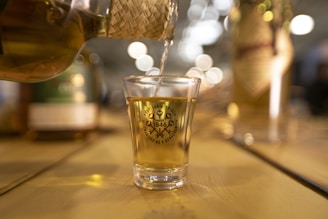 A shot glass filled with a golden liquid is being poured from a woven-covered bottle. The glass is placed on a wooden surface and has a logo printed on it. In the background, there are blurred lights and additional bottles that create a warm, inviting atmosphere.