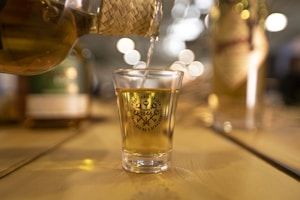 A shot glass filled with a golden liquid is being poured from a woven-covered bottle. The glass is placed on a wooden surface and has a logo printed on it. In the background, there are blurred lights and additional bottles that create a warm, inviting atmosphere.