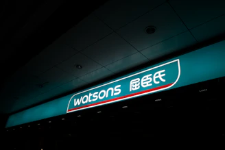 A illuminated storefront sign displaying the logo and name 'Watsons' in both English and Chinese characters. The sign is prominently situated on a teal background with a sleek design.
