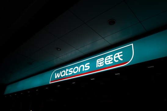 A illuminated storefront sign displaying the logo and name 'Watsons' in both English and Chinese characters. The sign is prominently situated on a teal background with a sleek design.