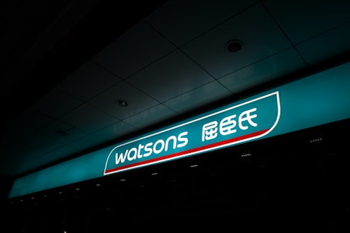 A illuminated storefront sign displaying the logo and name 'Watsons' in both English and Chinese characters. The sign is prominently situated on a teal background with a sleek design.