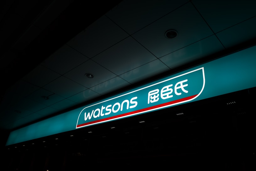 A illuminated storefront sign displaying the logo and name 'Watsons' in both English and Chinese characters. The sign is prominently situated on a teal background with a sleek design.