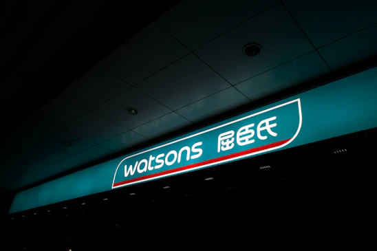 A illuminated storefront sign displaying the logo and name 'Watsons' in both English and Chinese characters. The sign is prominently situated on a teal background with a sleek design.