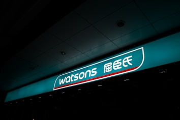 A illuminated storefront sign displaying the logo and name 'Watsons' in both English and Chinese characters. The sign is prominently situated on a teal background with a sleek design.