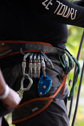A person is wearing a climbing harness with several carabiners and a belay device attached. The equipment is secured around their waist, and the harness is black with orange accents. The visible hand appears to be adjusting or holding one of the carabiners. There's a logo on the harness, and a rope is visible in the background.