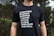 A person is wearing a black t-shirt with white text listing various regions and countries including Guatemala, Ecuador, Australia, England, Mexico, Zambia, Middle East, and North Africa. The background consists of greenery which seems to indicate an outdoor setting.