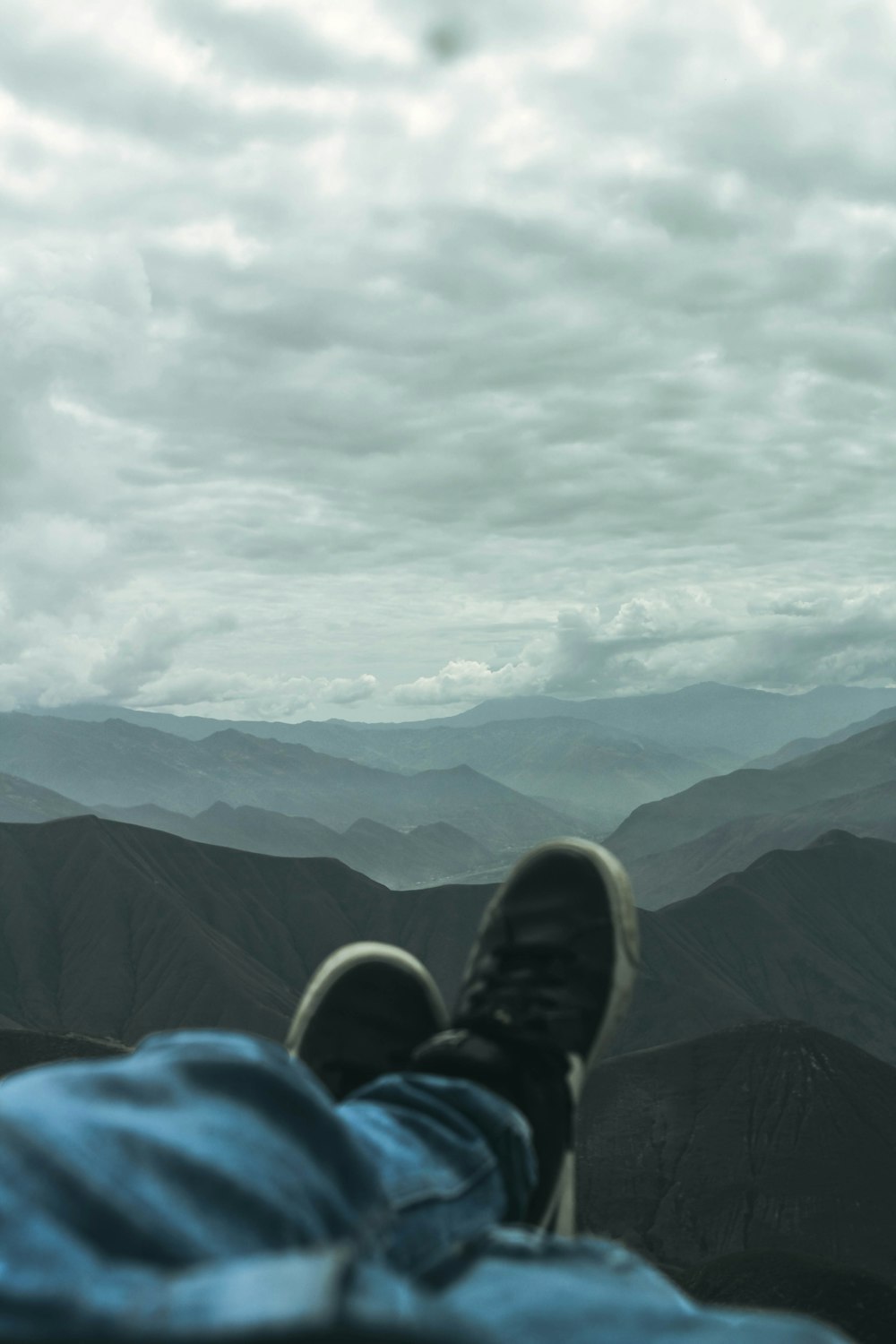 person in blue denim jeans and black hiking shoes sitting on rock mountain under white clouds