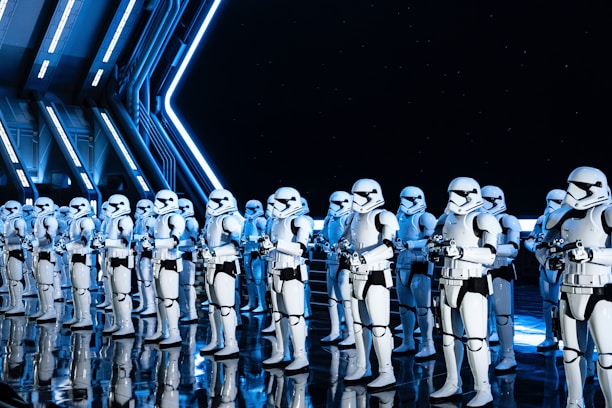 A group of uniformed characters dressed in white armored suits with black accents, resembling futuristic soldiers, stand in formation inside a high-tech, industrial-looking environment. The scene is illuminated by blue and white lighting, reflecting off the sleek, shiny floor and metal walls.