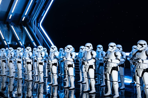 A group of uniformed characters dressed in white armored suits with black accents, resembling futuristic soldiers, stand in formation inside a high-tech, industrial-looking environment. The scene is illuminated by blue and white lighting, reflecting off the sleek, shiny floor and metal walls.