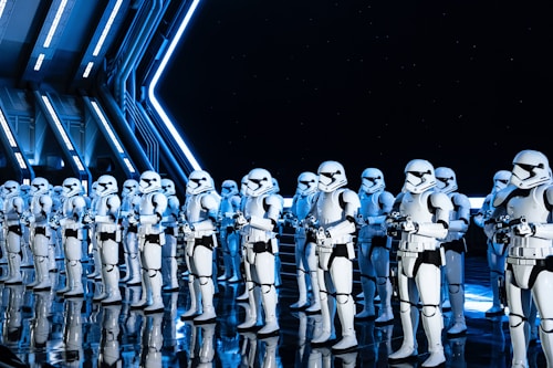 A group of uniformed characters dressed in white armored suits with black accents, resembling futuristic soldiers, stand in formation inside a high-tech, industrial-looking environment. The scene is illuminated by blue and white lighting, reflecting off the sleek, shiny floor and metal walls.