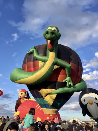 A vibrant scene featuring several colorful hot air balloons in the shape of characters. The central balloon resembles a green dinosaur with outstretched arms, set against a clear sky. Surrounding it are balloons in the shape of a pirate and a penguin. A large crowd of people is gathered below, looking upwards, creating a lively and festive atmosphere.