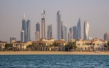 A vibrant cityscape featuring a row of luxury beachside villas in the foreground and a skyline of modern, high-rise buildings with distinct architectural designs in the background. The scene appears tranquil, with blue water in the foreground and clear skies above.