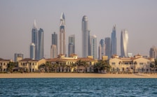 A vibrant cityscape featuring a row of luxury beachside villas in the foreground and a skyline of modern, high-rise buildings with distinct architectural designs in the background. The scene appears tranquil, with blue water in the foreground and clear skies above.