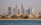 A vibrant cityscape featuring a row of luxury beachside villas in the foreground and a skyline of modern, high-rise buildings with distinct architectural designs in the background. The scene appears tranquil, with blue water in the foreground and clear skies above.