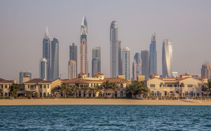 A vibrant cityscape featuring a row of luxury beachside villas in the foreground and a skyline of modern, high-rise buildings with distinct architectural designs in the background. The scene appears tranquil, with blue water in the foreground and clear skies above.