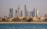 A vibrant cityscape featuring a row of luxury beachside villas in the foreground and a skyline of modern, high-rise buildings with distinct architectural designs in the background. The scene appears tranquil, with blue water in the foreground and clear skies above.
