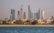 A vibrant cityscape featuring a row of luxury beachside villas in the foreground and a skyline of modern, high-rise buildings with distinct architectural designs in the background. The scene appears tranquil, with blue water in the foreground and clear skies above.