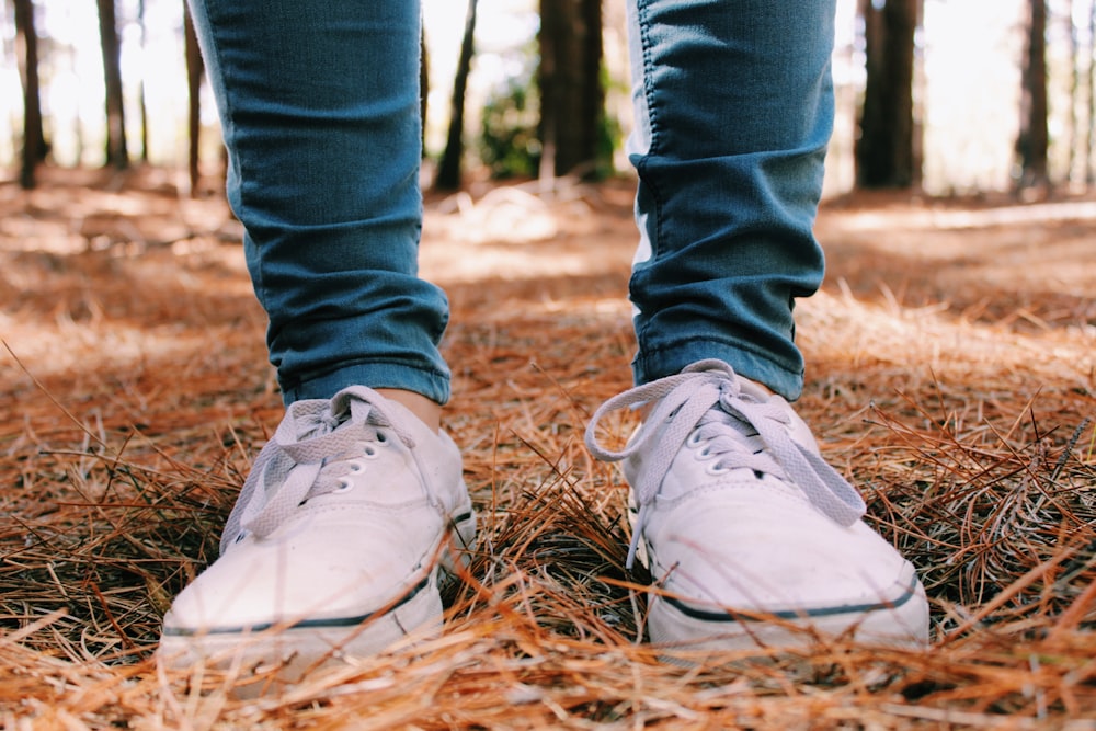 Persona con jeans de mezclilla azules y zapatillas blancas de pie sobre hojas secas