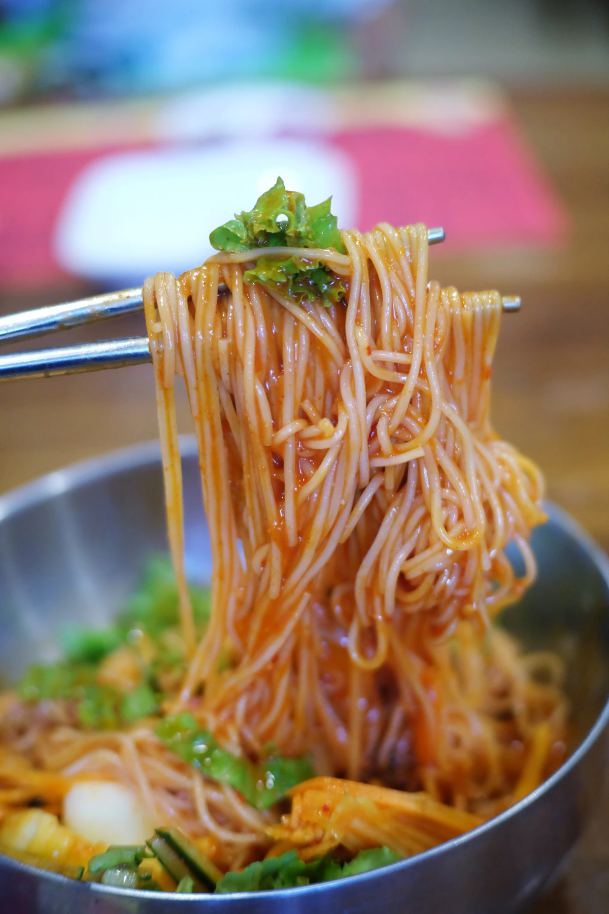 A bowl of spicy Korean cold noodles being lifted with chopsticks