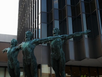 Three bronze statues of human figures with arms outstretched stand in front of a modern, glass-paneled building. The statues are artistically posed, conveying a sense of movement and unity.