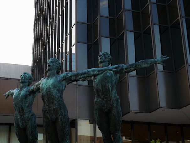 Three bronze statues of human figures with arms outstretched stand in front of a modern, glass-paneled building. The statues are artistically posed, conveying a sense of movement and unity.