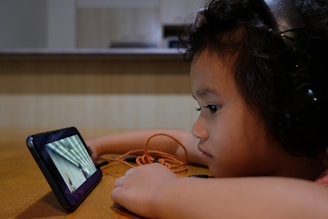 A child wearing headphones is intently watching a video on a smartphone placed on a table. The child has curly hair and appears focused on the screen, with an orange cable also visible. The background is a blurred room setting, suggesting an indoor environment.