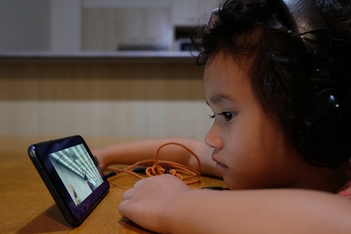 A child wearing headphones is intently watching a video on a smartphone placed on a table. The child has curly hair and appears focused on the screen, with an orange cable also visible. The background is a blurred room setting, suggesting an indoor environment.