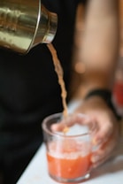 A beverage is being poured from a metal shaker into a transparent glass held by a hand. The liquid appears to be orange in color, and there is some foam forming at the top.
