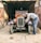 An older man is working on a vintage car in a dilapidated garage. The car has a rusty orange body with a vintage number plate reading 'WBA 288'. The surroundings are gritty and worn, featuring a metallic garage door and a dusty floor.