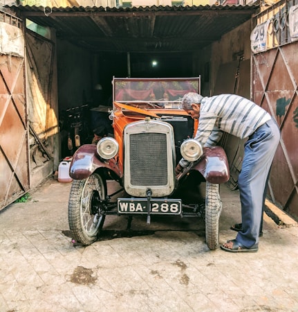 An older man is working on a vintage car in a dilapidated garage. The car has a rusty orange body with a vintage number plate reading 'WBA 288'. The surroundings are gritty and worn, featuring a metallic garage door and a dusty floor.