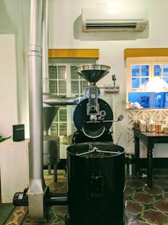 A coffee roasting machine is placed in an indoor setting, with an air conditioning unit on the wall above it. The machine is large and industrial-looking, with a glass window on the front, allowing a view of the roasting process. Next to it, there is a vent or pipe leading upwards. A window with white shutters is behind the machine. To the right, a wooden table holds various packaged items and a small bin is placed underneath.