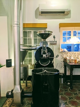 A coffee roasting machine is placed in an indoor setting, with an air conditioning unit on the wall above it. The machine is large and industrial-looking, with a glass window on the front, allowing a view of the roasting process. Next to it, there is a vent or pipe leading upwards. A window with white shutters is behind the machine. To the right, a wooden table holds various packaged items and a small bin is placed underneath.