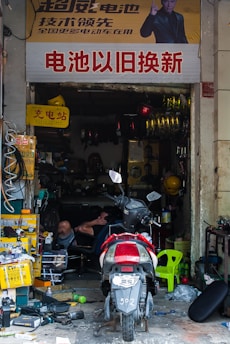 A cluttered repair shop with a motorcycle parked in front. The shop is filled with various tools, boxes, and equipment. A person is reclining on a chair inside, surrounded by spare parts and machinery. A large sign with Chinese characters is displayed above the entrance.
